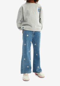 Felpa grigia con grafica di margherite, abbinata a jeans a zampa d'elefante blu con ricami bianchi di margherite e sneakers chiare.