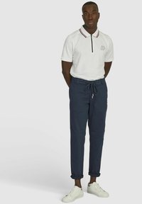 Weißes Polohemd mit Reißverschluss und roten sowie marineblauen Akzenten, kombiniert mit marineblauen Jogginghosen und weißen Turnschuhen. Einfache, lässige Gestaltung.