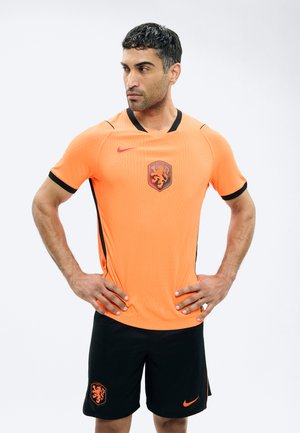 Man met Nederlands voetbalshirt in oranje en zwarte korte broek, handen in de zij, kijkt zijwaarts tegen een effen witte achtergrond.