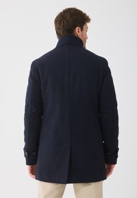 Mies, der einen navyblauen Wollmantel mit hohem Kragen und Knopfmanschetten trägt, steht mit dem Rücken zu einem schlichten weißen Hintergrund.