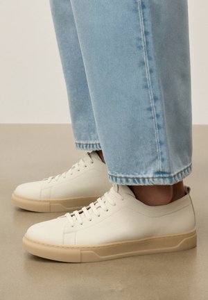 Bílé nízké kožené tenisky s béžovými podrážkami, nošené s světlými modrými džínami, stojící na hladkém béžovém povrchu.