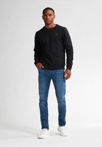 Zwart sweatshirt met een ronde hals, voorzien van een klein bruin label, gecombineerd met blauwe jeans en witte sneakers, staande op een effen achtergrond.