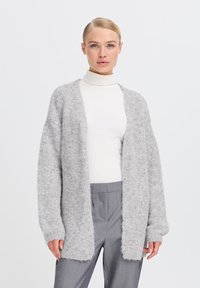 Cardigan grigio chiaro fluffy con apertura frontale, maniche lunghe e polsini a costine, indossato su un dolcevita bianco e pantaloni grigi.