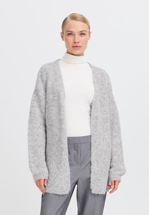 Cardigan grigio chiaro fluffy con apertura frontale, maniche lunghe e polsini a costine, indossato su un dolcevita bianco e pantaloni grigi.