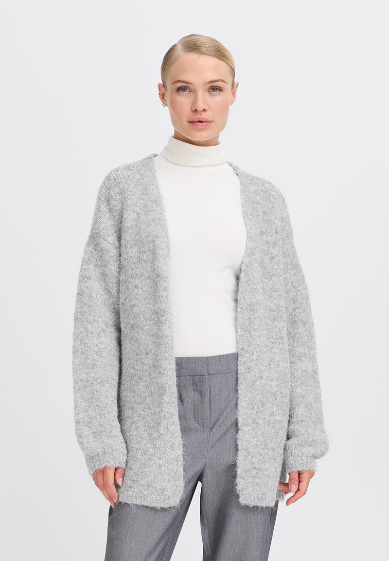 Cardigan grigio chiaro fluffy con apertura frontale, maniche lunghe e polsini a costine, indossato su un dolcevita bianco e pantaloni grigi.