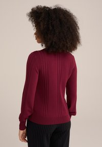 Bordó pulovr s žebrovanými detaily, přiléhavými manžetami a texturovaným vzorem na zádech. Vyrobený z měkkého pleteného materiálu.