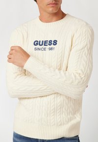 Pulover alb din tricot cu model imitat de cablu, având pe piept brodat în albastru marin "GUESS SINCE 1981"; guler rotund și mansete și tiv cu model rib. 