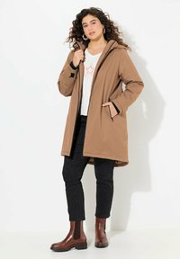 Manteau marron à capuche avec fermeture éclair à l'avant, poches latérales et bordures noires, associé à un pantalon à motifs sombres et des bottines marron.