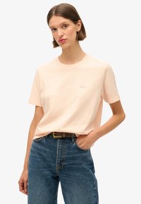 T-shirt en coton pêche clair avec un col rond, des manches courtes et un logo brodé subtil. Associé à un jean en denim bleu taille haute.