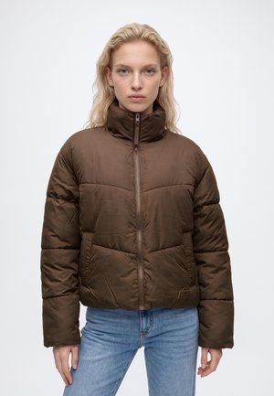 Jeune femme aux longs cheveux blonds porte une doudoune marron zippée, associée à un jean bleu clair, debout devant un fond blanc uni.
