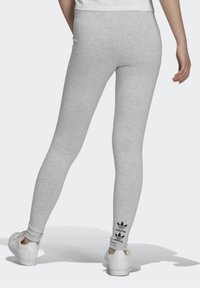 Person som bär ljusgrå Adidas leggings och vita sneakers, står med ett ben något böjt, Adidas-logotypen nära fotleden.