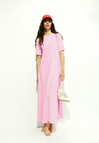 Pink, short-sleeved maxi dress with a relaxed fit. Model holds white perforated footwear. Background is plain white.
