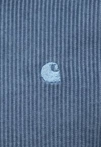 Modrá kordurová látka s jemným modrým vyšitou logom. Povrch má vertikálne priehlbiny a logo je umiestnené v dolnej časti.