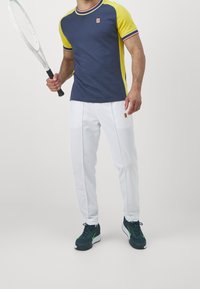 Námořnické a žluté sportovní tričko s krátkým rukávem a červenými akcenty, spárované s bílými kalhotami s logem a tmavými teniskami. Drží tenisovou raketu.