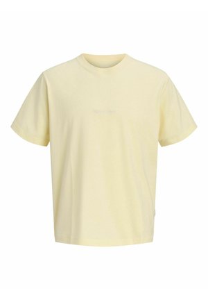 Garçon blond debout, portant un T-shirt jaune pâle, un short en jean longueur genoux, des chaussettes blanches et des chaussures beiges à lacets, regardant vers la droite.