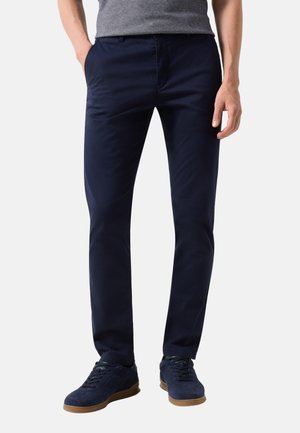 Mężczyzna noszący granatowe wąskie spodnie i pasujące granatowe tenisówki z gumowymi podeszwami, stojący z ręką w kieszeni.