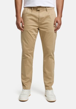 Hombre con pantalones beige de corte recto y zapatillas blancas, de pie con las manos relajadas a los lados sobre fondo blanco.