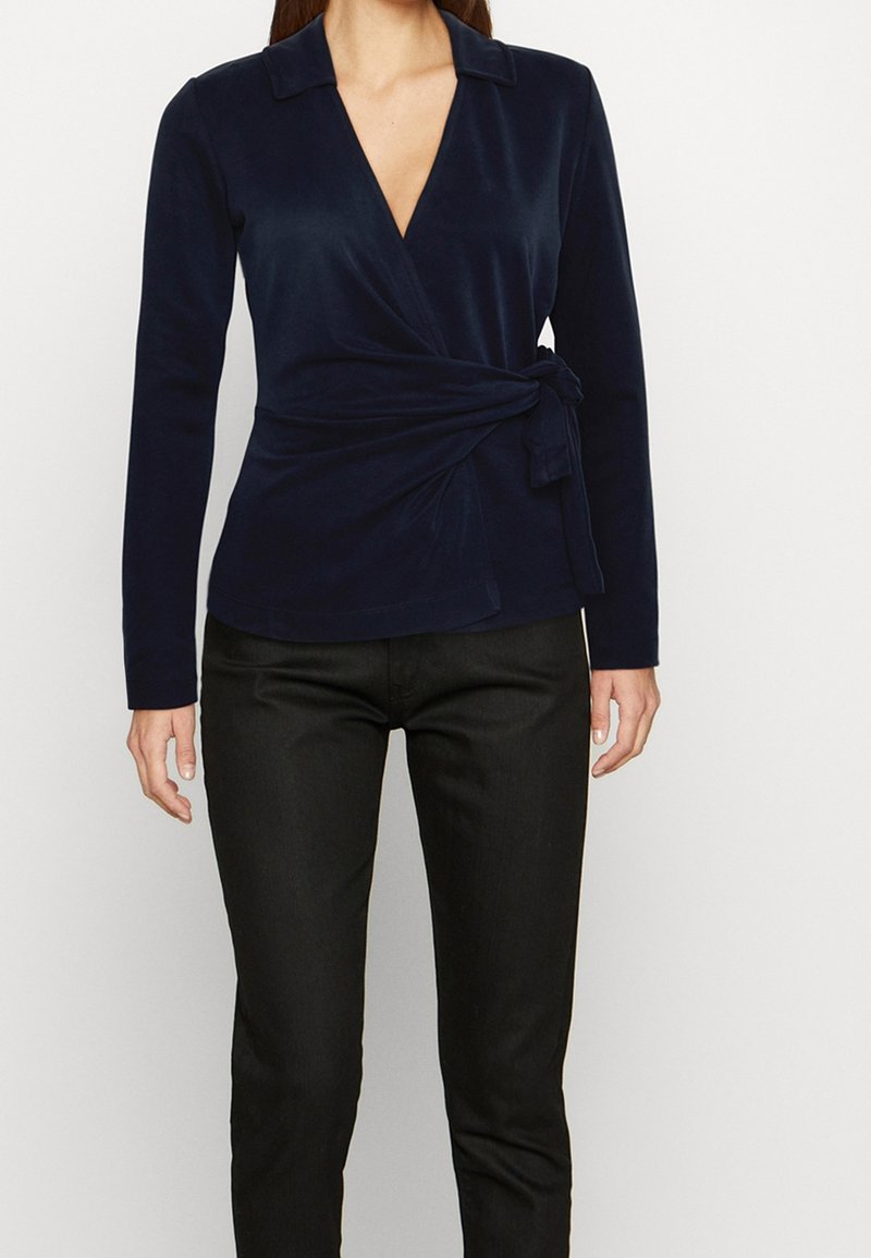 Femme portant un chemisier cache-cœur bleu marine ajusté à manches longues et un pantalon noir slim sur fond blanc uni.