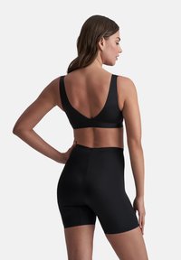 Donna vista di spalle che indossa un reggiseno sportivo nero e pantaloni da ciclismo neri a vita alta su uno sfondo bianco uniforme.
