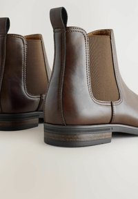 Botas Chelsea de cuero marrón con lados elásticos, mostrando un primer plano de los detalles de costura y el tacón contra un fondo claro.