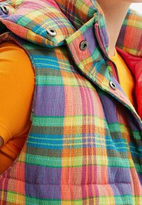 Karierte Weste in lebhaften Farben (Pink, Grün, Blau, Orange) mit metallischen Druckknöpfen und einem hohen Kragen; über einem orangen Hemd getragen.