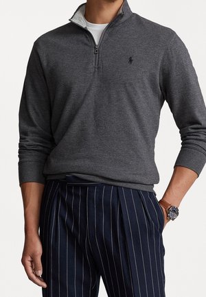 Mężczyzna w ciemnoszarym swetrze zapinanym do ćwiartki na zamek, na białej koszuli, granatowych spodniach w prążki oraz z brązowym zegarkiem na skórzanym pasku na nadgarstku.