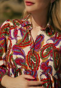 Bunte Bluse mit Paisleymuster in Creme, verziert mit roten, grünen und lila Akzenten. Knopfverschluss vorne, kragen und geraffte Taille.