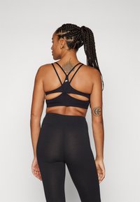 Crni sportski crop top s dizajnom s trakama na leđima i izrezanim detaljima. Upareno s crnim tajicama visokog struka, prikazano s leđa.