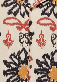 Chemise à manches courtes en tissu léger avec des motifs floraux multicolores en rouge, marine et jaune sur fond crème. Six boutons visibles.