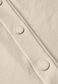 Beige stof met een gladde textuur heeft drie ronde, platte knopen en gestikte naden, die de constructiedetails van het kledingstuk benadrukken.