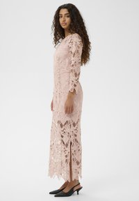 Rochie lungă roz din dantelă cu modele florale, mâneci trei sferturi și fante laterale. Purtați cu tocuri slingback negre, cu vârful ascuțit.