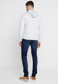 Sweat à capuche gris avec cordon de serrage, poignets et ourlet côtelés ; associé à un jean bleu foncé et des baskets beiges. Détail de couture visible sur le jean.