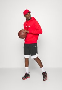 Červená mikina s kapucňou a logom býka, čierne kraťasy s bielymi akcentmi, čierno-červené tenisky, drží basketbal. Jednoduché pozadie.