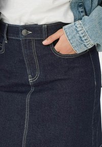 Dżinsowa spódnica o ciemnoniebieskim wykończeniu, z kontrastowymi szwami, przednią kieszenią i metalowym guzikiem w pasie.