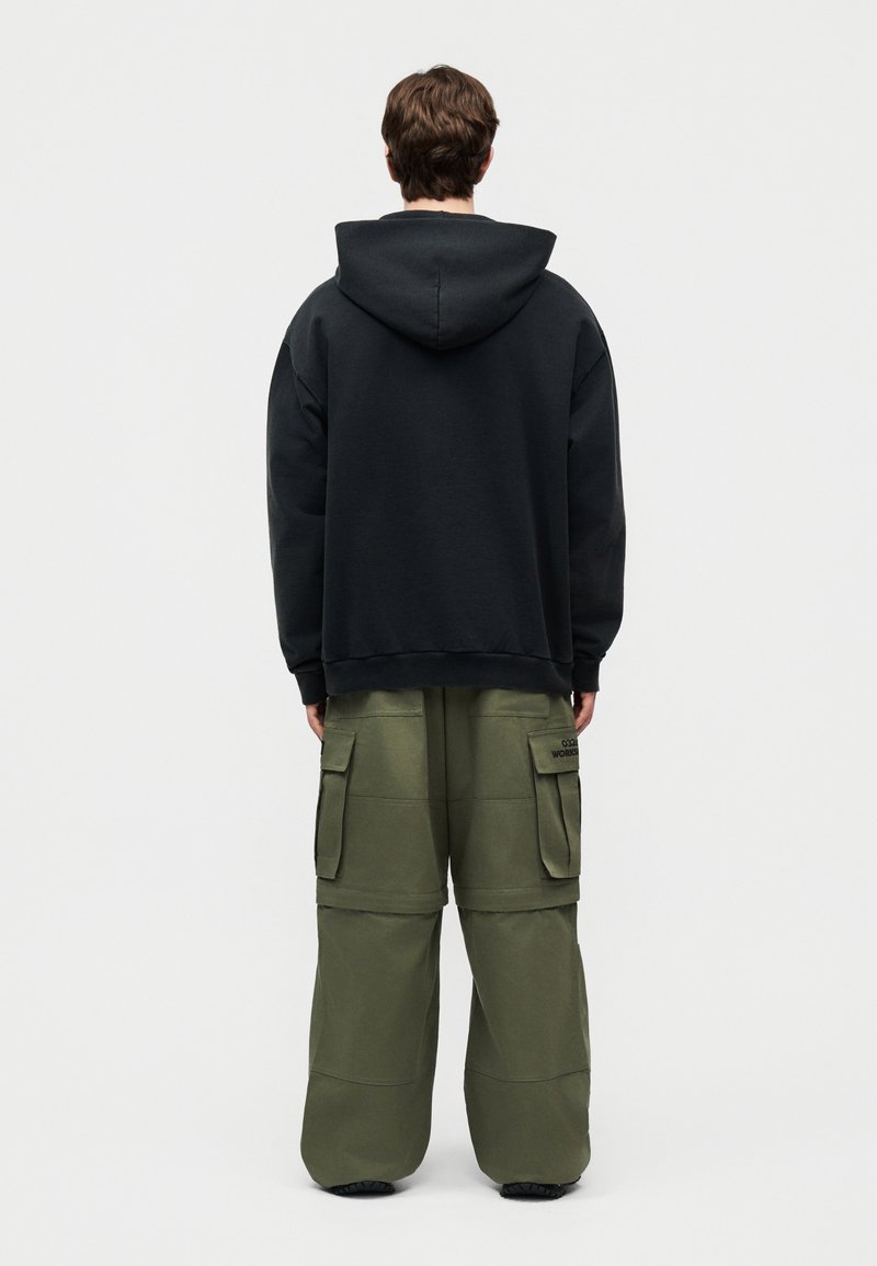 Persoon draagt een zwart sweatshirt met capuchon en loszittende olijfgroene cargobroek, staand met de rug naar voren op een eenvoudige lichte achtergrond.
