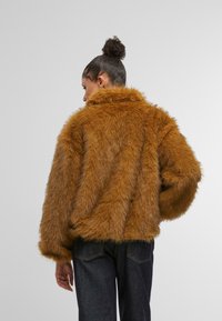 Sieviete ar tumšiem matiem, kas sasieti bunā, ģērbusies brūnā pūkainā mākslīgā kažokā un tumšās džinsās, attēlota no aizmugures uz neitrāla fona.