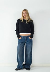 Zwarte cropped trui, lichte tanktop eronder, gecombineerd met wijde blauwe jeans met voorzakken en een denim textuur. Zwarte schoenen.