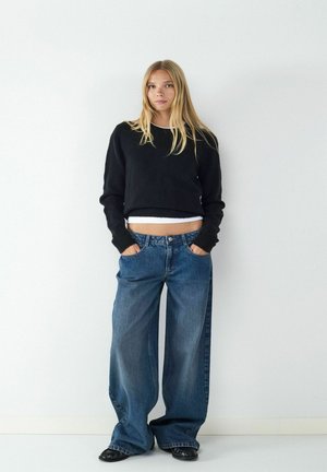 Zwarte cropped trui, lichte tanktop eronder, gecombineerd met wijde blauwe jeans met voorzakken en een denim textuur. Zwarte schoenen.
