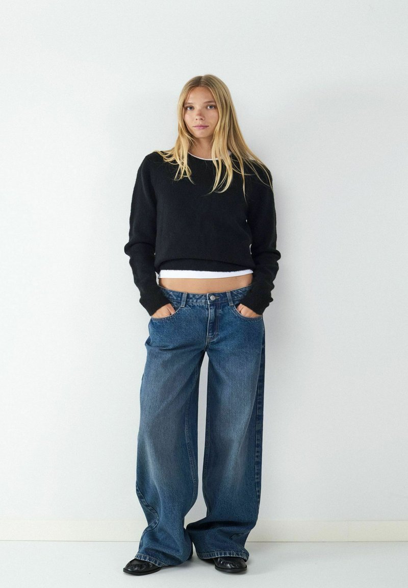 Zwarte cropped trui, lichte tanktop eronder, gecombineerd met wijde blauwe jeans met voorzakken en een denim textuur. Zwarte schoenen.