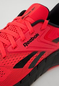 Fényes vörös Reebok futócipő fekete részletekkel. Hálós felsőrész, lapos fűzők, struktúrált talp, és logó a nyelvén és az oldalán.