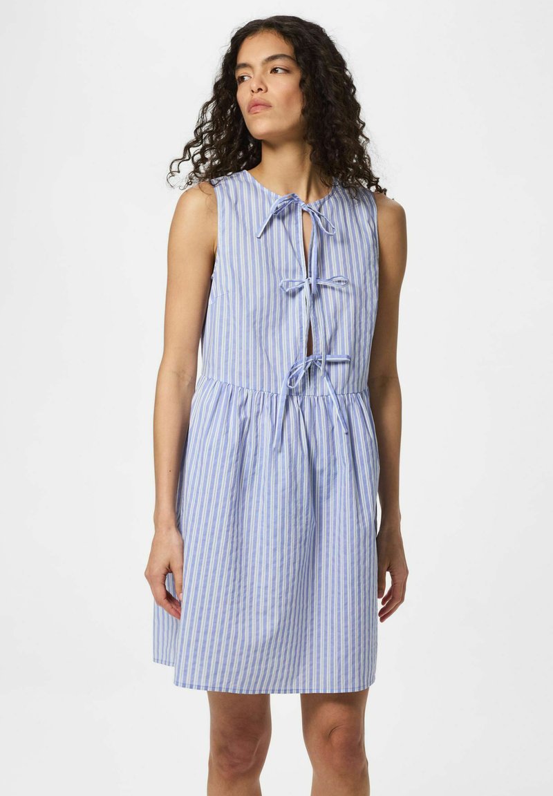 Femme aux cheveux bouclés portant une robe sans manches bleu et blanc à rayures verticales avec trois fermetures à nouer sur le devant.