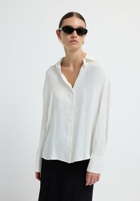 Femme portant des lunettes de soleil noires, des boucles d'oreilles en or, une chemise blanche boutonnée ample et une jupe plissée noire, se tenant devant un fond blanc uni.