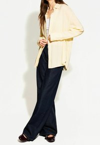 Geel gestreept overhemd met knopen, relaxte pasvorm, gecombineerd met donkere brede broek en donkere schoenen. Soepele textuur, lange mouwen en hoge halslijn.