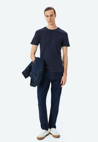 Marineblå ribbet t-shirt parret med mørkeblå denimjeans og hvide sneakers. Modellen holder en denimjakke, hvilket fremviser et afslappet, lagdelt look.