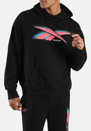 Man wearing black Reebok hoodie and sweatpants with large red and blue Reebok logo on chest and smaller logo on left thigh.