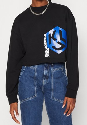Person iført sort cropped sweatshirt med blåt Karl Lagerfeld Jeans-logo og højtaljede blå jeans, accessoriseret med sølvkædehalskæde.