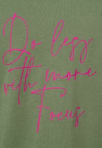 Olivgrön tyg med en mjuk textur som har rosa kursiv text: "Gör mindre med mer fokus" tydligt placerad i mitten.