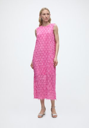 Naine, kes kannab varrukateta roosat midi kleiti tekstuursete ümmarguste mustritega ja hõbedasi avatud varbaga plokkkontsaga sandaale, seistes ühevärvilise valge tausta ees.