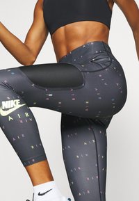 Atletka v černých legínách Nike Air s multikolorovým textem a síťovanou kapsou, v bílých ponožkách Nike, zvedající koleno v cvičební pozici.