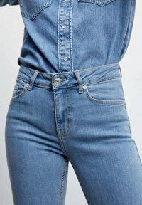 Gros plan sur une personne portant un jean denim bleu clair et une chemise en denim assortie rentrée à l'intérieur, sur un fond uni.